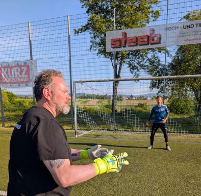 Ein Torwart hält den Ball, während ein Spieler vor dem Tor steht.