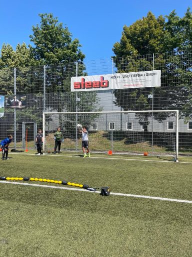 Kinder trainieren im Fußballtor auf einem Rasenplatz, umgeben von Bäumen.