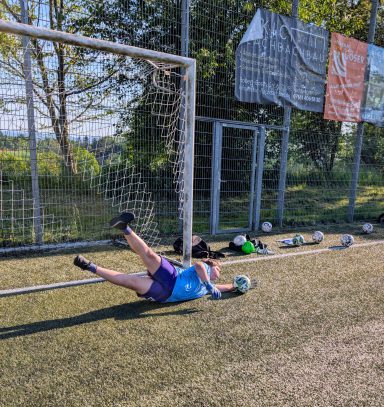 Torwart hält einen Schuss im Training, liegt auf dem Rasen mit Händen am Ball.