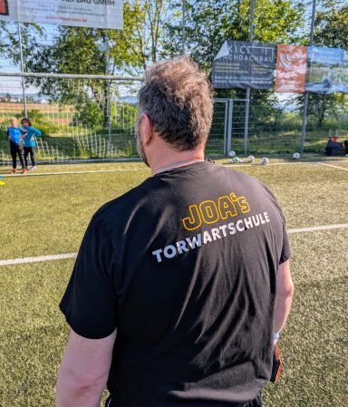 Person mit kurzem Haar und schwarzem Shirt, auf steht auf einem Fußballplatz.