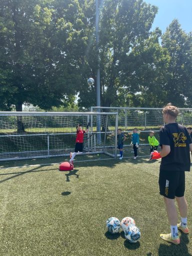 Kinder üben Fußballschießen auf einem Rasenplatz unter blauem Himmel.