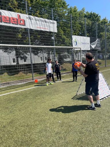 Zwei Kinder vor einem Tor, während ein Erwachsener ihnen beim Fußballspielem hilft.