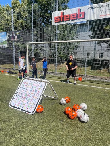 Zwei Personen im Tor, ein Torwart und ein Schütze, mit mehreren Bällen und einem Trainingsziel.