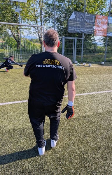 Person in schwarzem T-Shirt und Handschuhen steht auf einem Fußballfeld.
